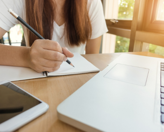 机の前にスマホとpcを置いてノートを取っている女性の画像