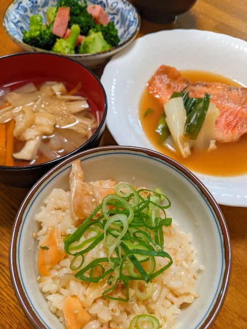 中央手前鮭の炊き込みご飯、左 汁物、右 赤魚の煮つけ、上 ブロッコリーとベーコンの炒め物の画像