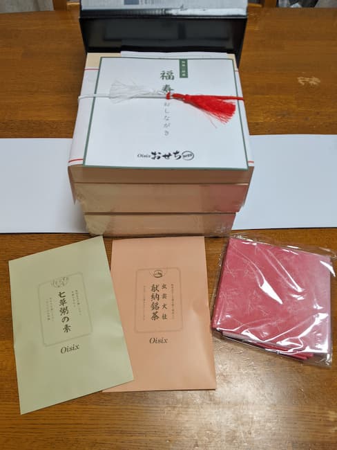 中央上 オイシックスおせち福寿、手前に封筒に入った七草粥の素・緑茶・ピンクの風呂敷の画像