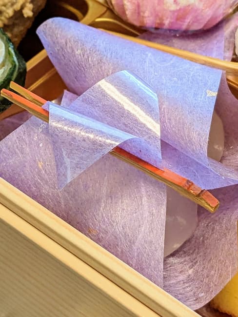 オイシックスおせち慶梅2023カスタード餅の画像。お餅が紫色の紙に包まれ、上を竹串で止められている